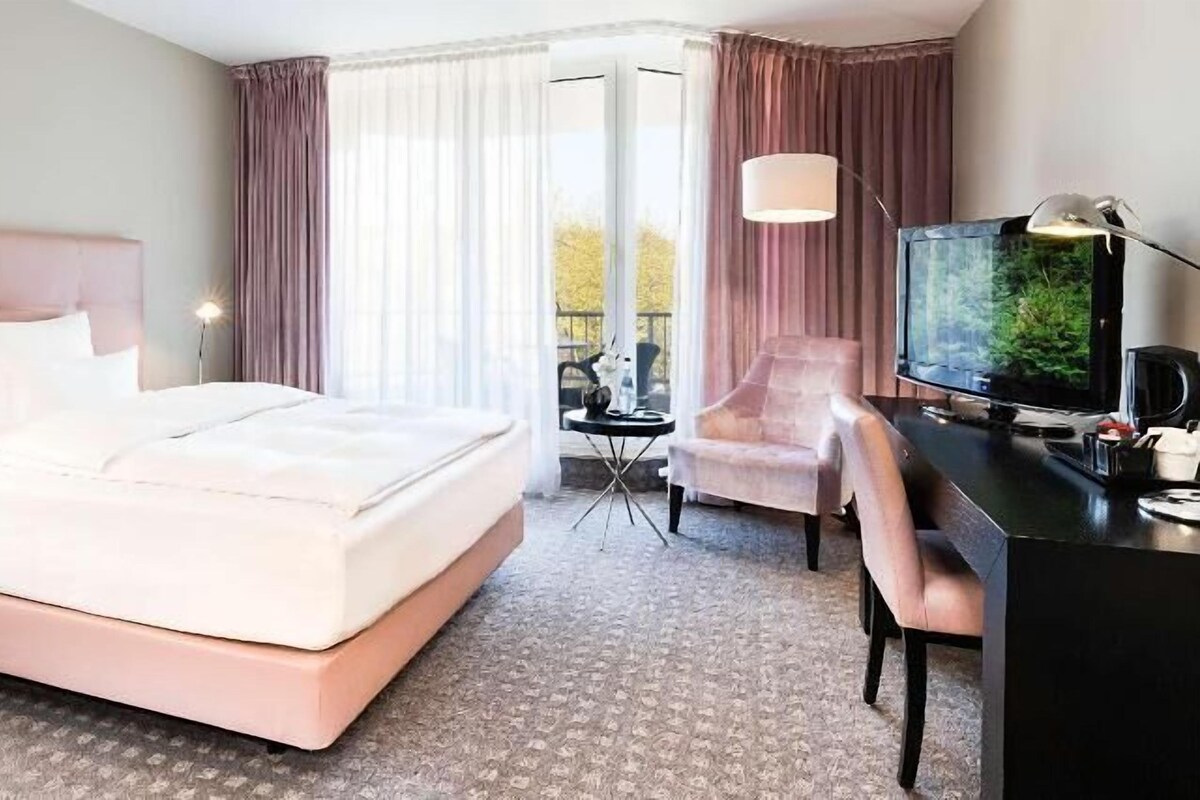 Una habitación de hotel con una cama grande, un escritorio con televisor, una silla y una mesita de noche.