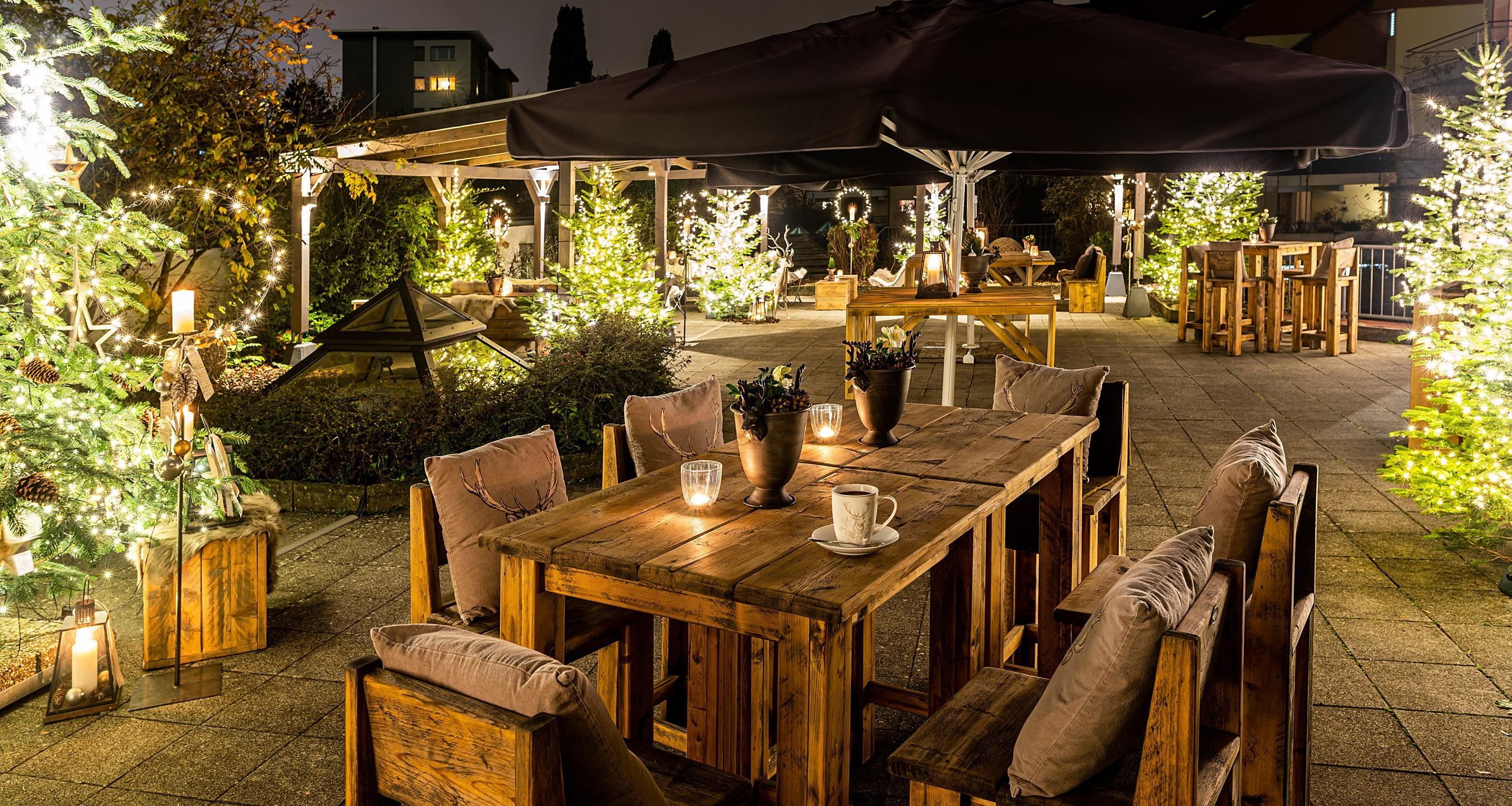 Een buiten eethoek met houten tafels en stoelen, omgeven door verlichte bomen en planten.