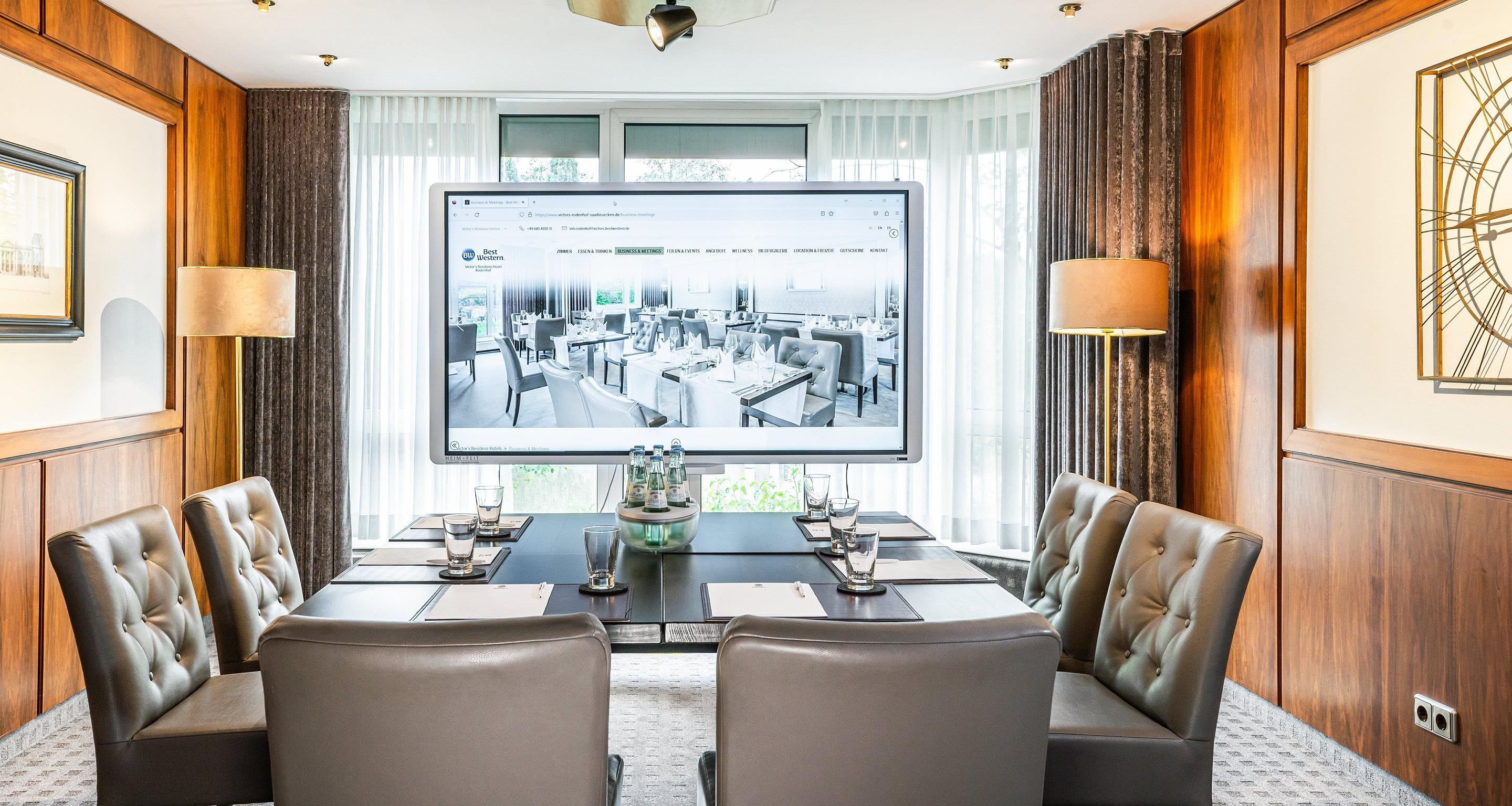Ein Konferenzraum mit einer großen Leinwand, die ein Hotelinnere zeigt, Ledersessel, einem Holztisch und Blick ins Freie durch große Fenster.