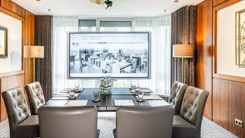 Ein Konferenzraum mit einer großen Leinwand, die ein Hotelinnere zeigt, Ledersessel, einem Holztisch und Blick ins Freie durch große Fenster.
