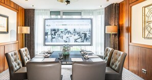 Un salón de conferencias con una pantalla grande que muestra el interior de un hotel, sillas de cuero, una mesa de madera y vistas al exterior a través de amplias ventanas.