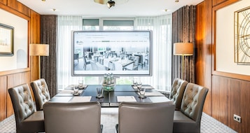 Un salón de conferencias con una pantalla grande que muestra el interior de un hotel, sillas de cuero, una mesa de madera y vistas al exterior a través de amplias ventanas.
