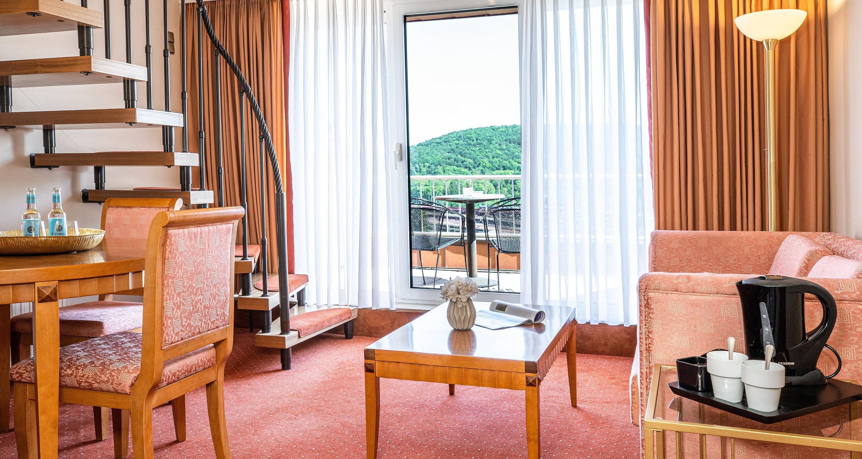 Ein Hotelzimmer mit einer Holztreppe, einem Essbereich mit Tisch und Stühlen, einem Sofa, einer Kaffeemaschine und Blick auf einen Balkon mit Gartenmöbeln.