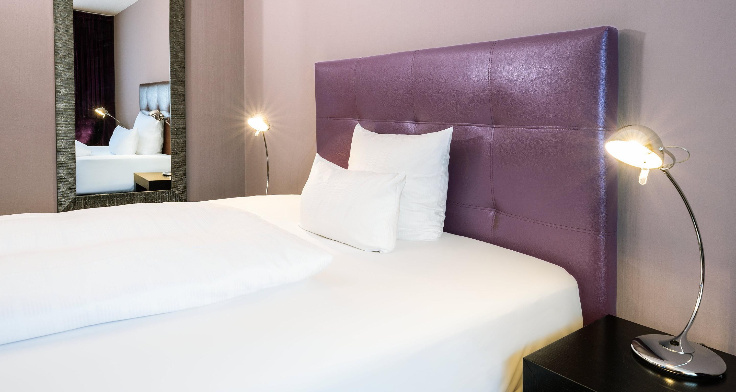 Ein ordentlich bezogenes Bett mit weißen Laken, einem violetten Kopfteil und zwei Nachttischlampen.