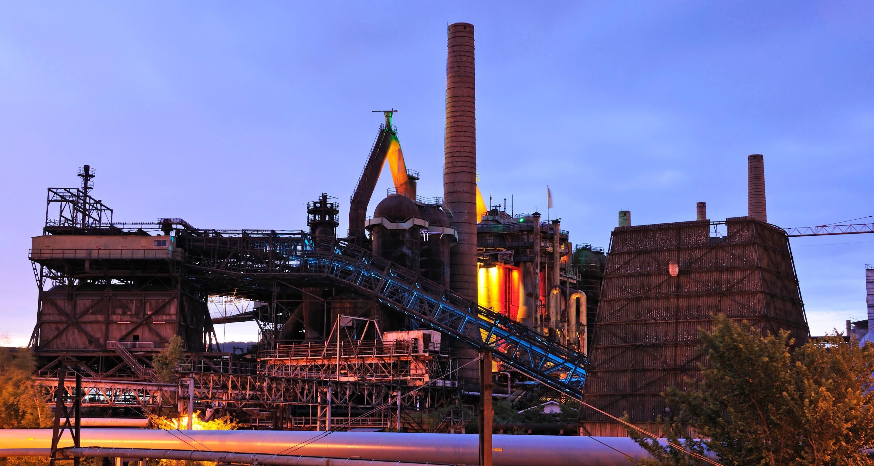 Un extenso complejo industrial con varias chimeneas altas, un sistema de cinta transportadora y una gran estructura cilíndrica.