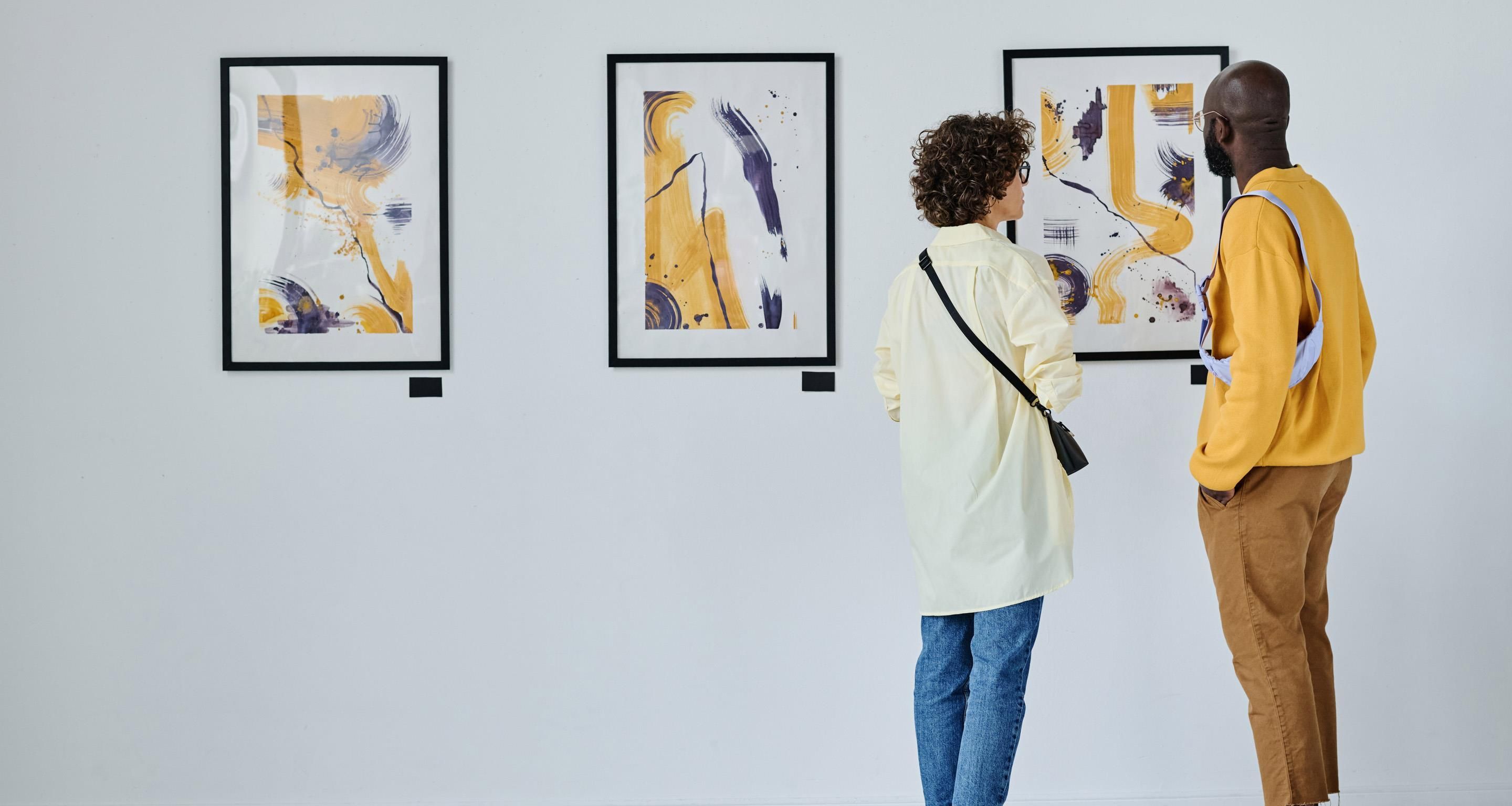 Two people examining framed artworks on a wall in a gallery.