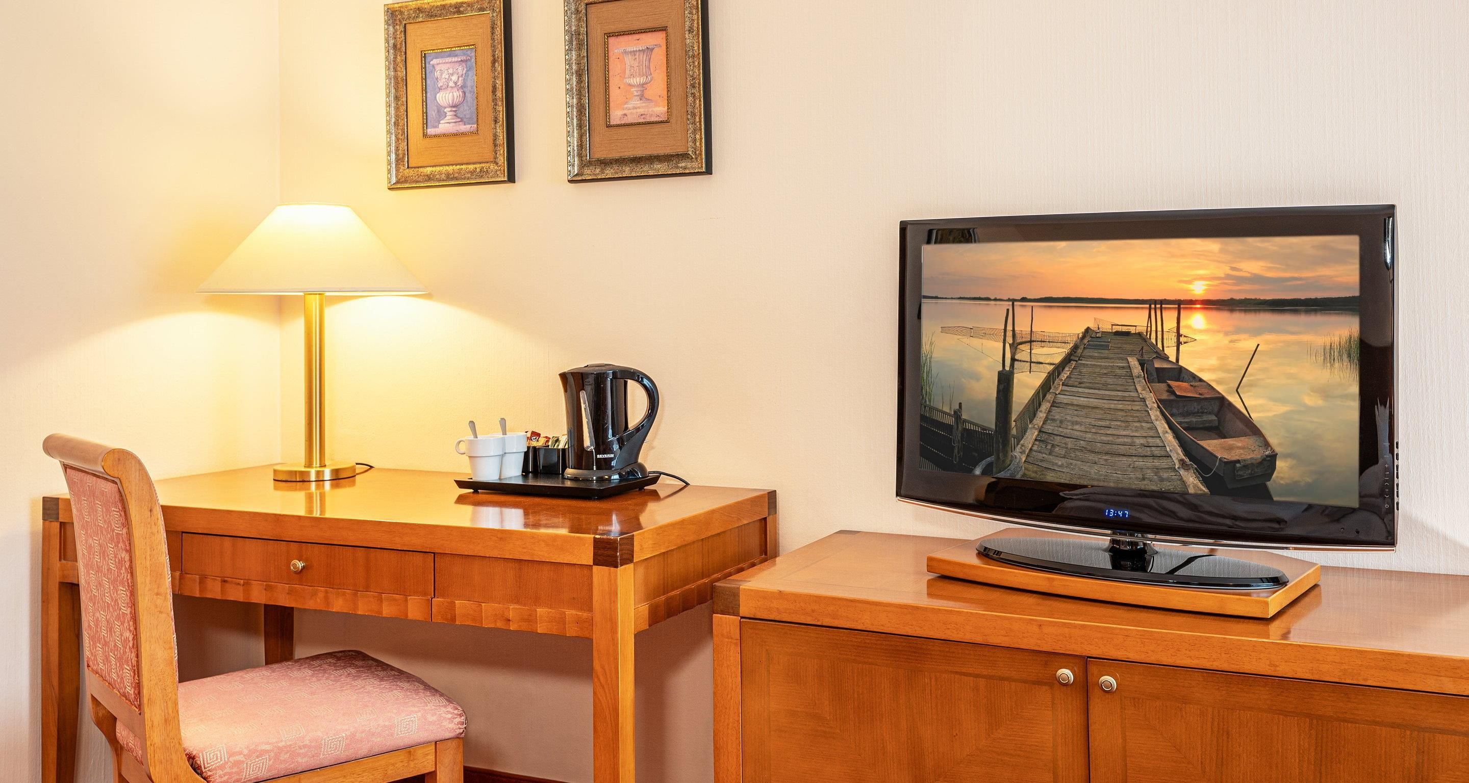 A wooden desk with a lamp, a chair, a coffee maker, and a television displaying a sunset scene.