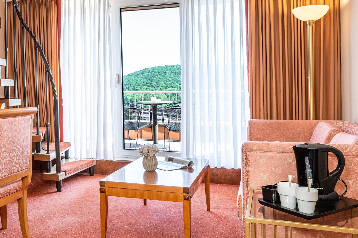 Habitación de hotel con una escalera de madera, un área de comedor con mesa y sillas, un sofá, cafetera y vistas a un balcón con mobiliario exterior.