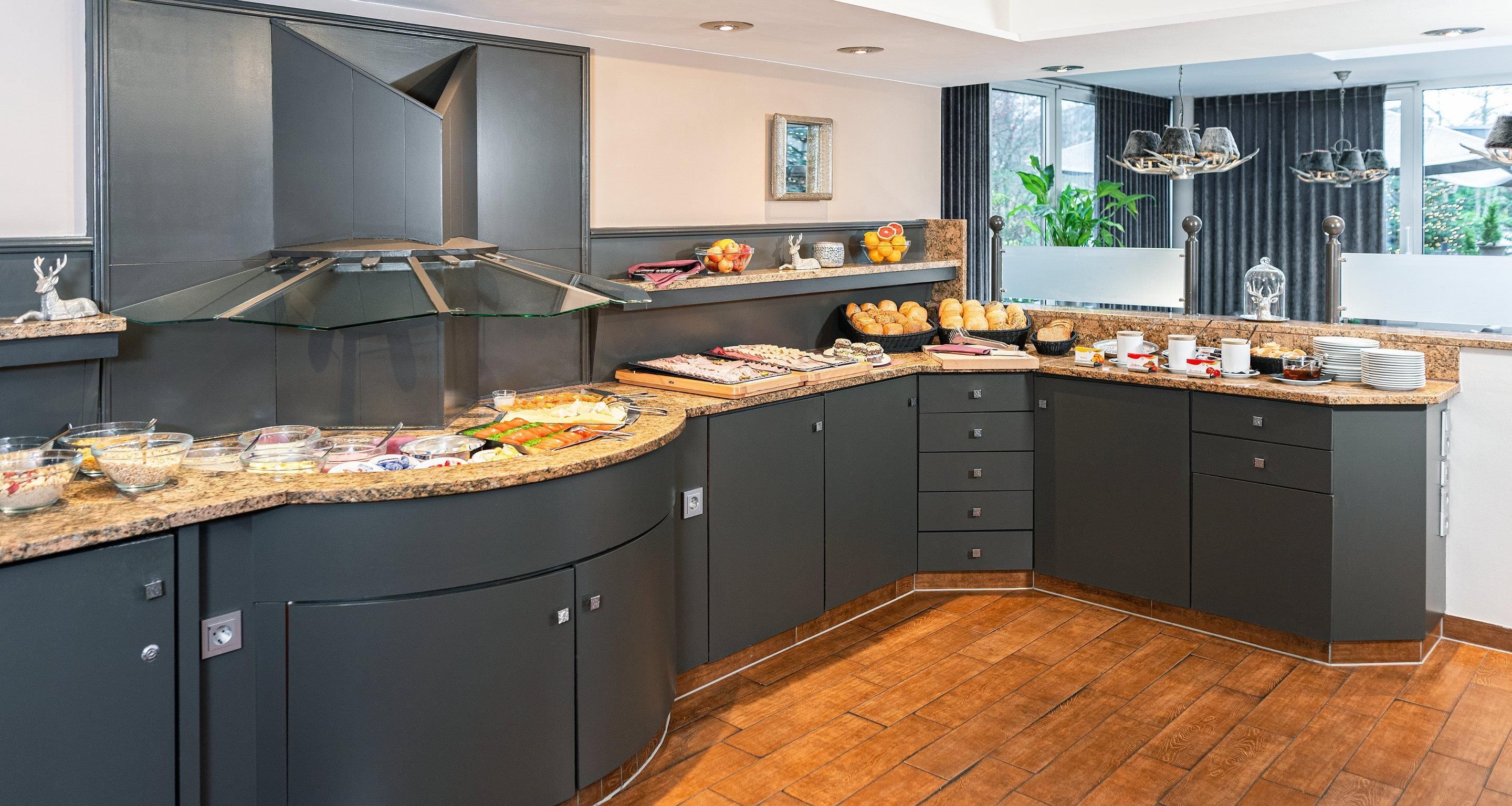 Une cuisine moderne avec des meubles de couleur sombre, un îlot central aux lignes courbes où sont disposés des éléments pour le petit-déjeuner, et une grande fenêtre offrant une vue sur la verdure.