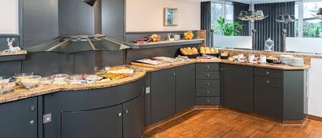A modern kitchen with dark cabinetry, a curved island with breakfast items, and a large window with a view of greenery.