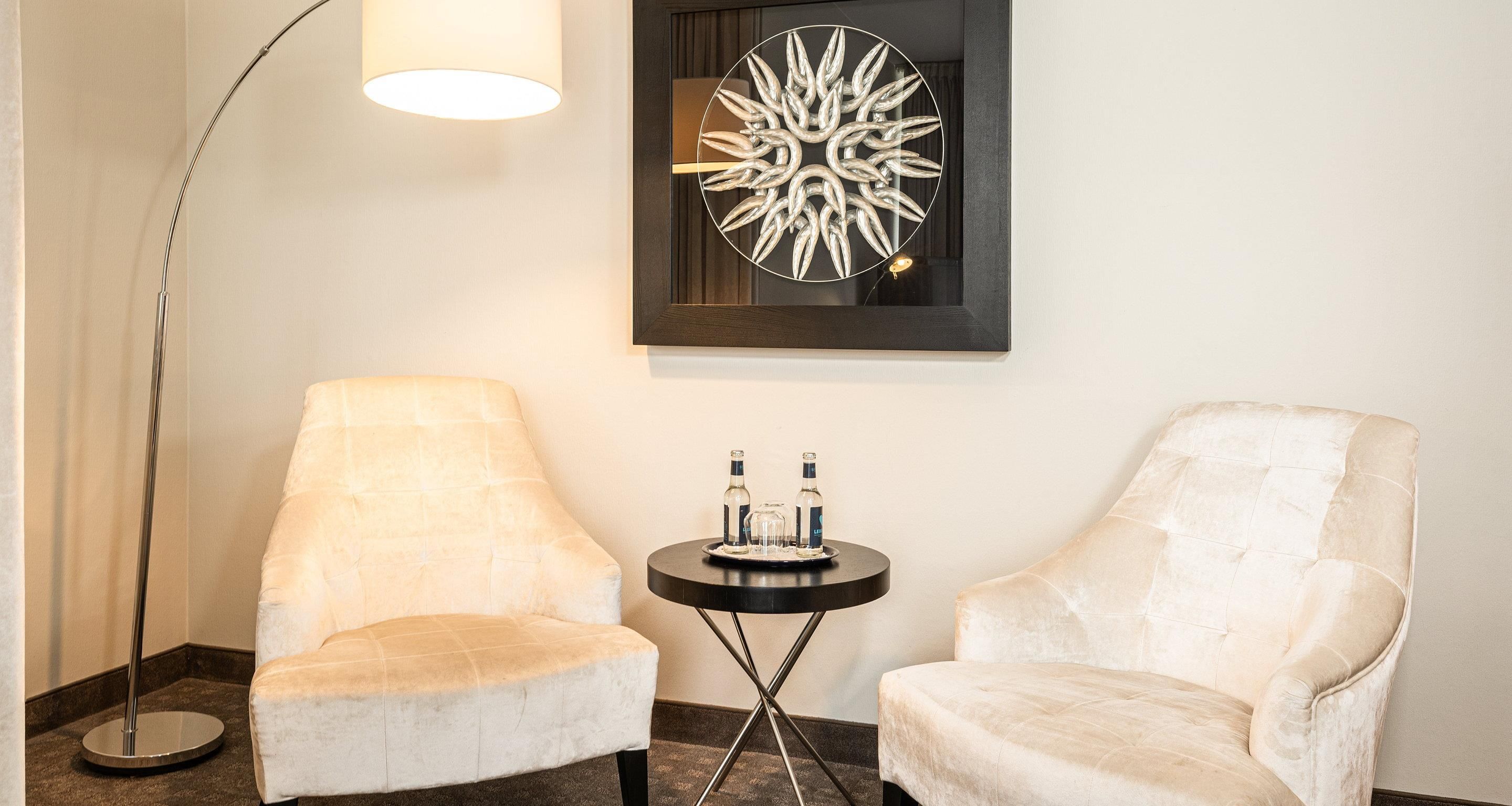A room with two white armchairs, a small round table with bottles, a floor lamp, and a wall art piece.