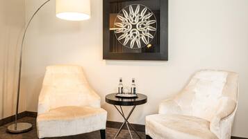 A room with two white armchairs, a small round table with bottles, a floor lamp, and a wall art piece.