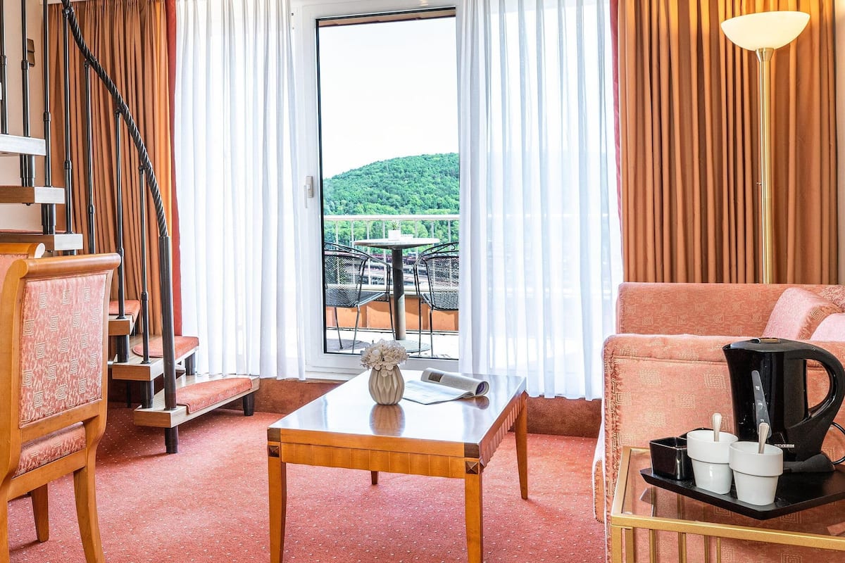 A hotel room with a wooden staircase, a dining area with a table and chairs, a sofa, a coffee maker, and a view of a balcony with outdoor furniture.