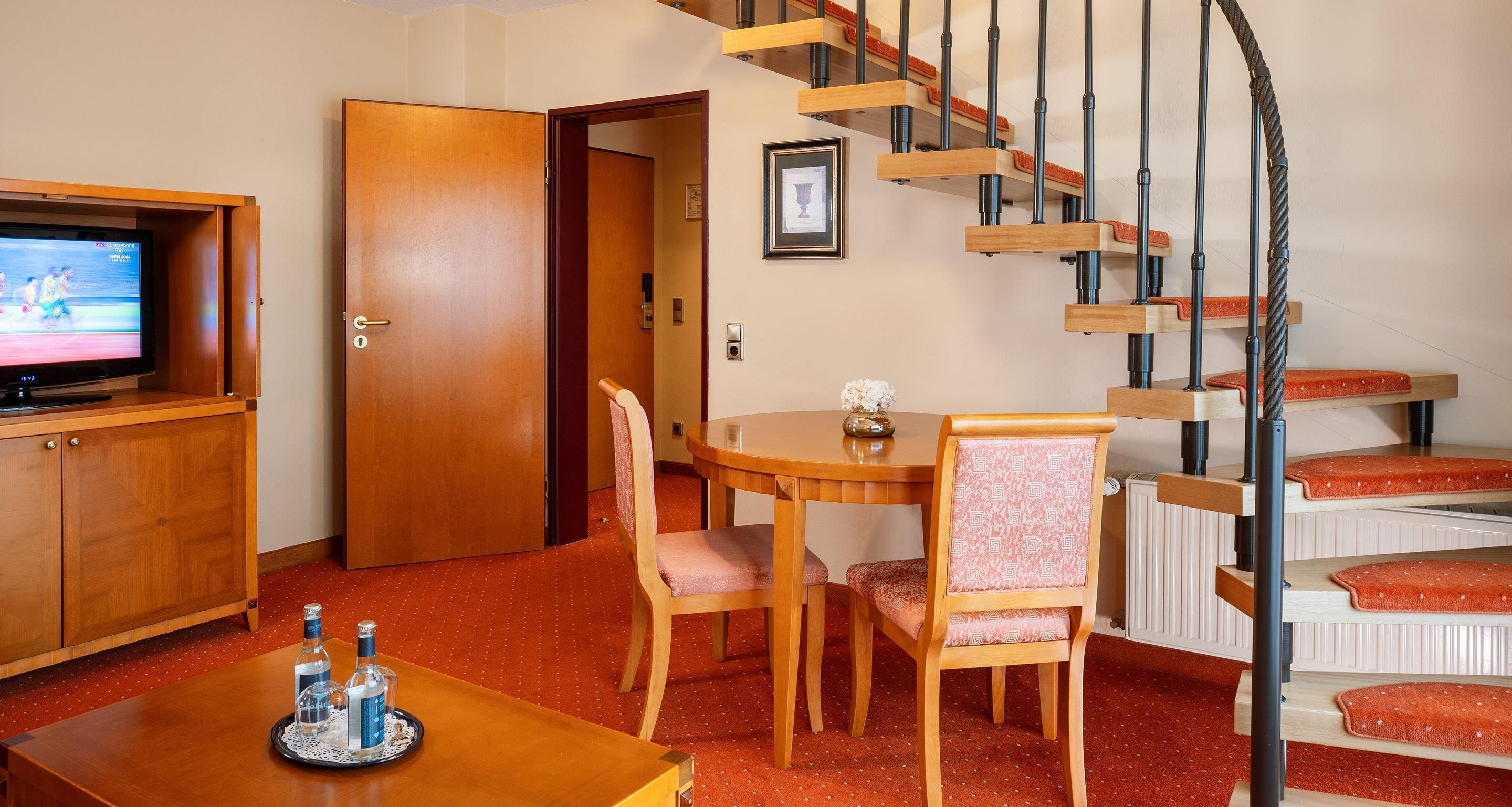 Ein Zimmer mit einem Holzes Esstisch, Stühlen, einem Fernseher, einer Treppe und einer Tür zu einem anderen Raum.