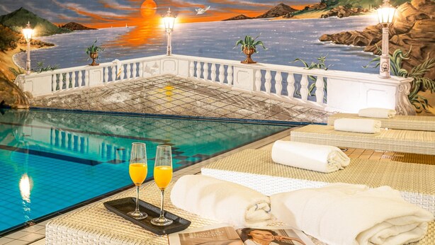 A pool area with a sunset mural, lounge chairs, towels, and two glasses of orange juice. - Best Western Victor's Residenz-Hotel Rodenhof (Saarbrücken)