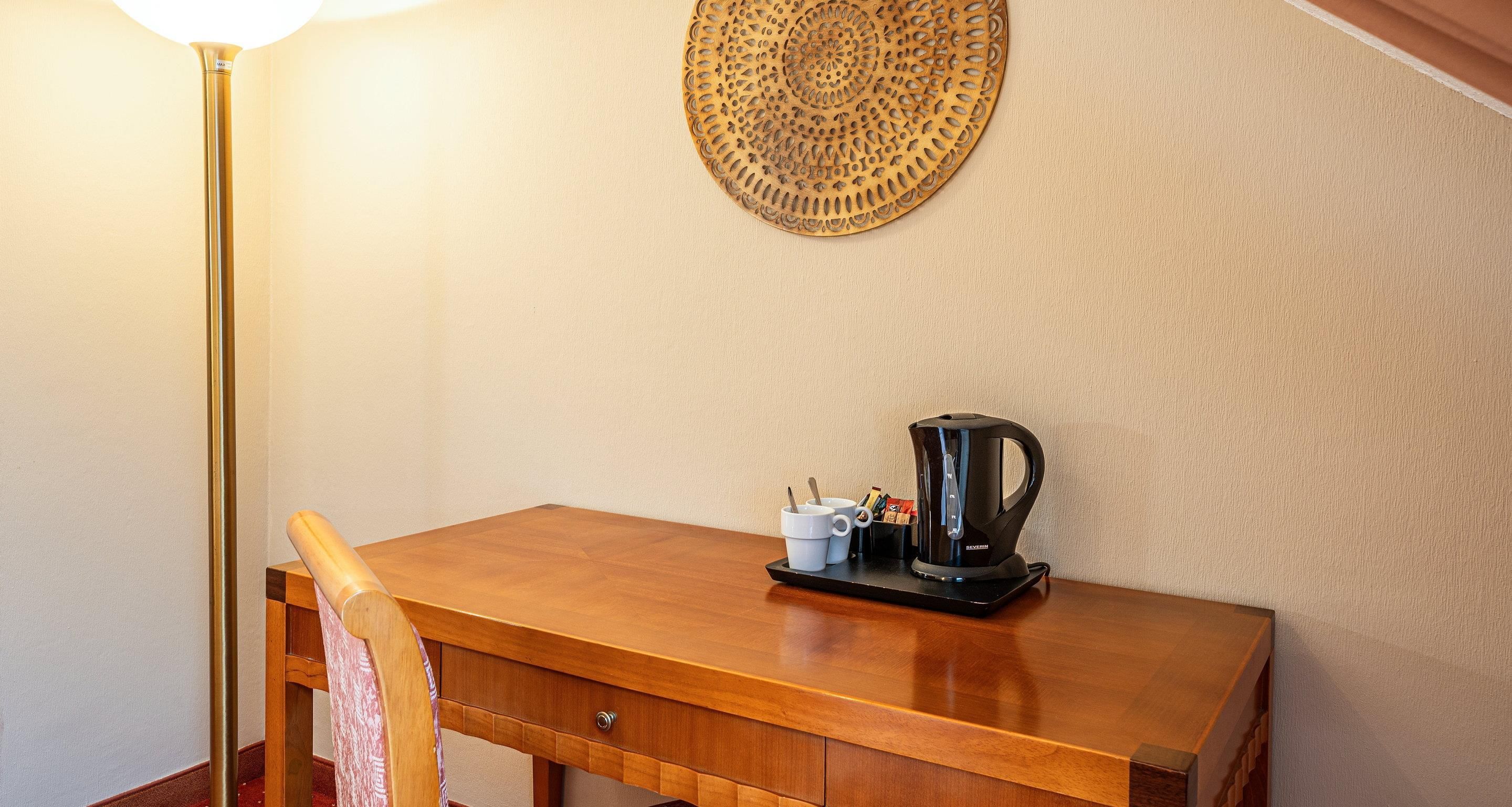 A wooden desk with a coffee maker, cups, and a tray, a chair, and a wall lamp.