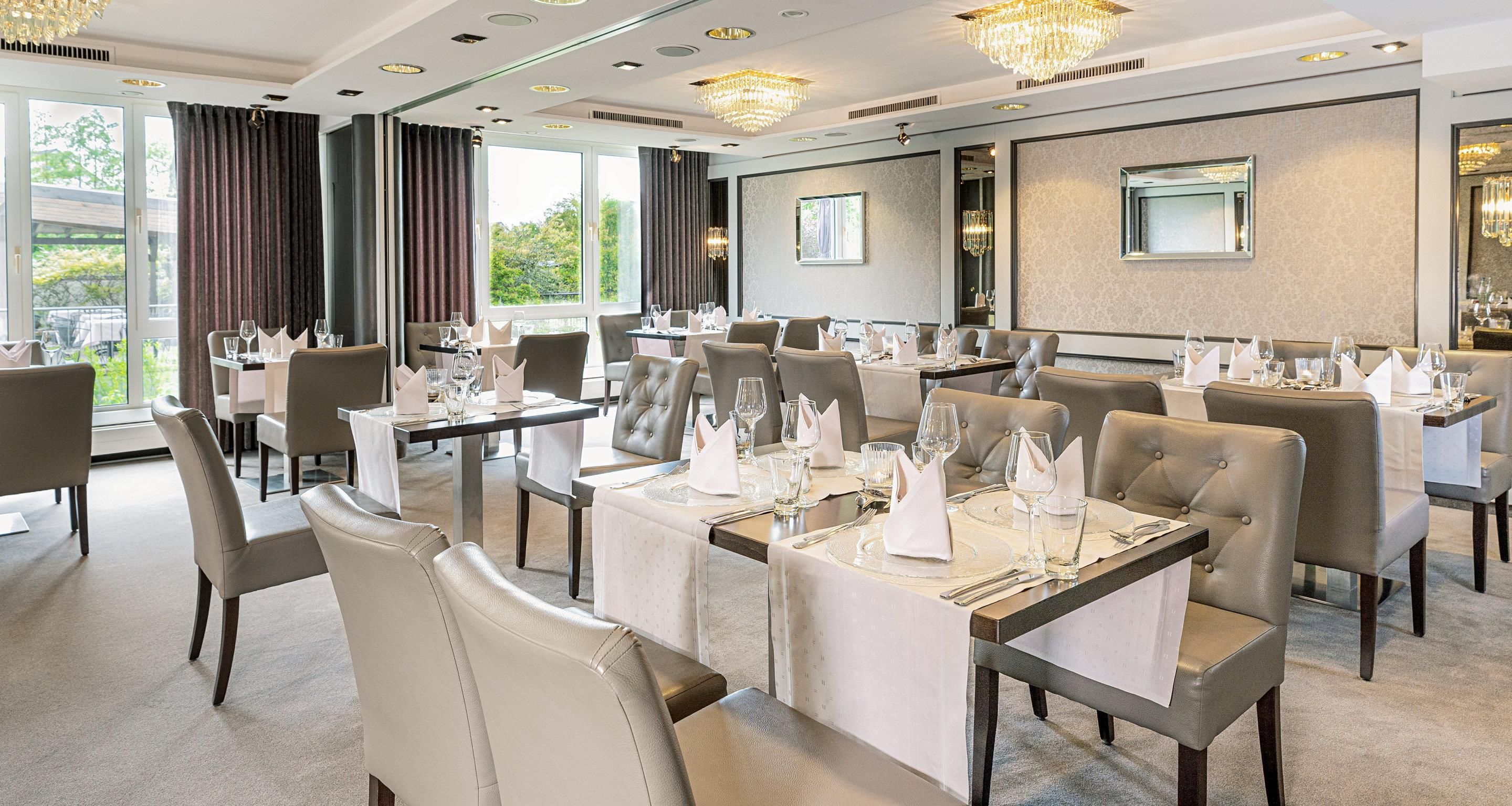 A dining area with tables set for service, large windows with curtains, and a chandelier.