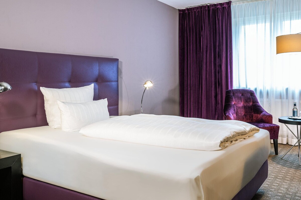 Habitación de hotel con una cama grande, un escritorio con lámpara, una silla y vistas al exterior a través de cortinas.