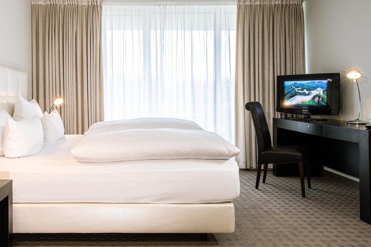 Una habitación de hotel con cama, escritorio, silla, televisor y cortinas.
