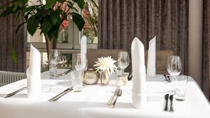 A table set with white napkins, silverware, and clear glasses, with a potted plant and decorative flowers in the background.