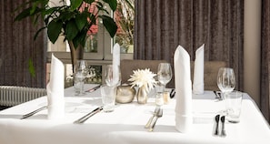 Una mesa puesta con servilletas blancas, cubiertos y vasos transparentes, con una planta en maceta y flores decorativas al fondo.
