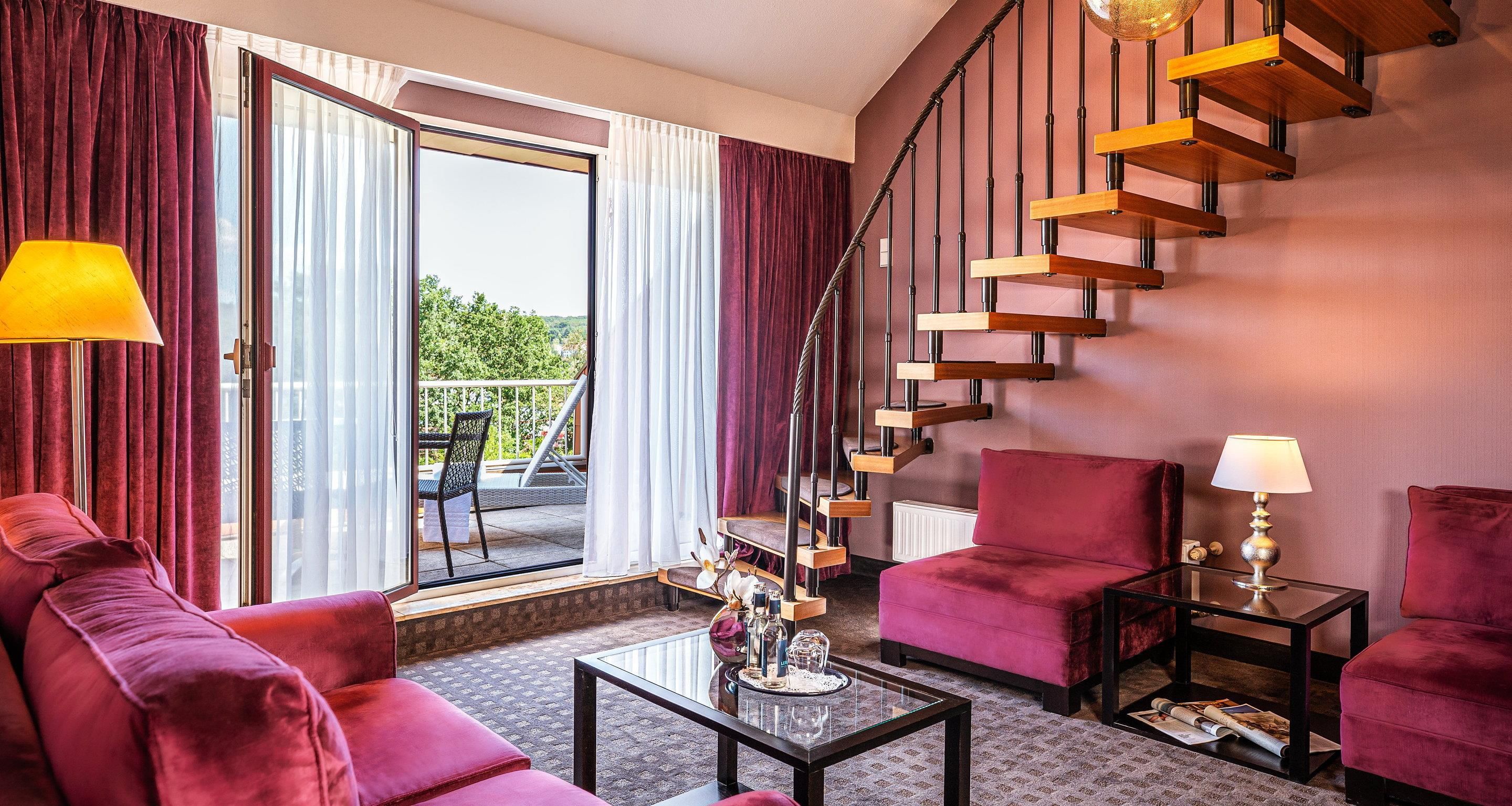 Una habitación de hotel moderna con un sofá rojo, una mesa de centro de vidrio, un sillón rojo, una pequeña mesa con una lámpara y una escalera de caracol.