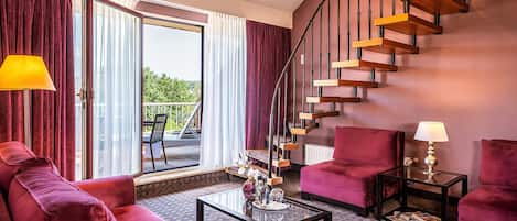 A modern hotel room with a red sofa, a glass coffee table, a red armchair, a small table with a lamp, and a spiral staircase.