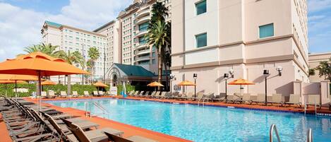 Area piscina di un hotel con lettini e ombrelloni, una piscina blu cristallina e un edificio a più piani sullo sfondo.