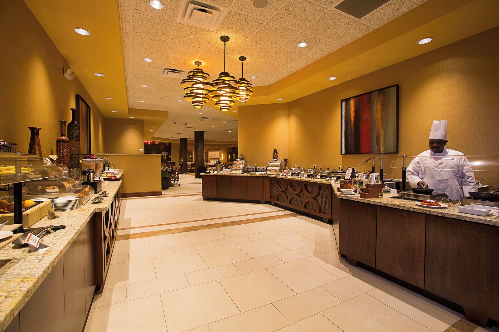 A hotel kitchen with a buffet setup, a chef preparing food, and a modern interior design.