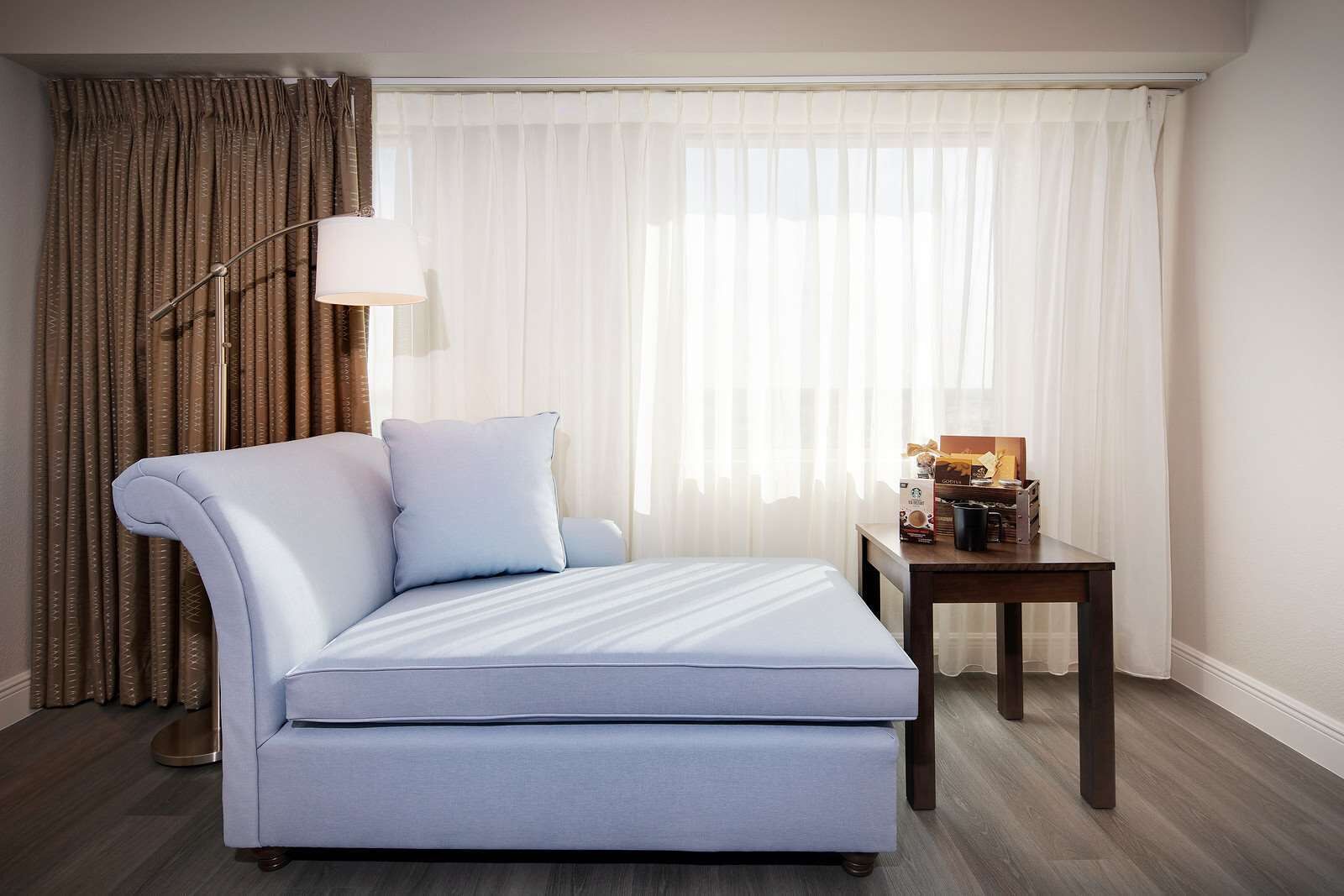 A hotel room with a bed, a side table with a lamp, and a view through sheer curtains.