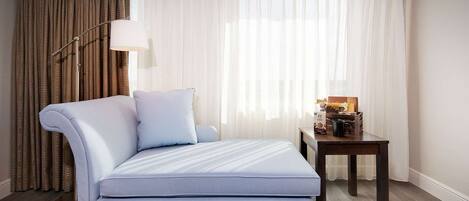 A hotel room with a bed, a side table with a lamp, and a view through sheer curtains.