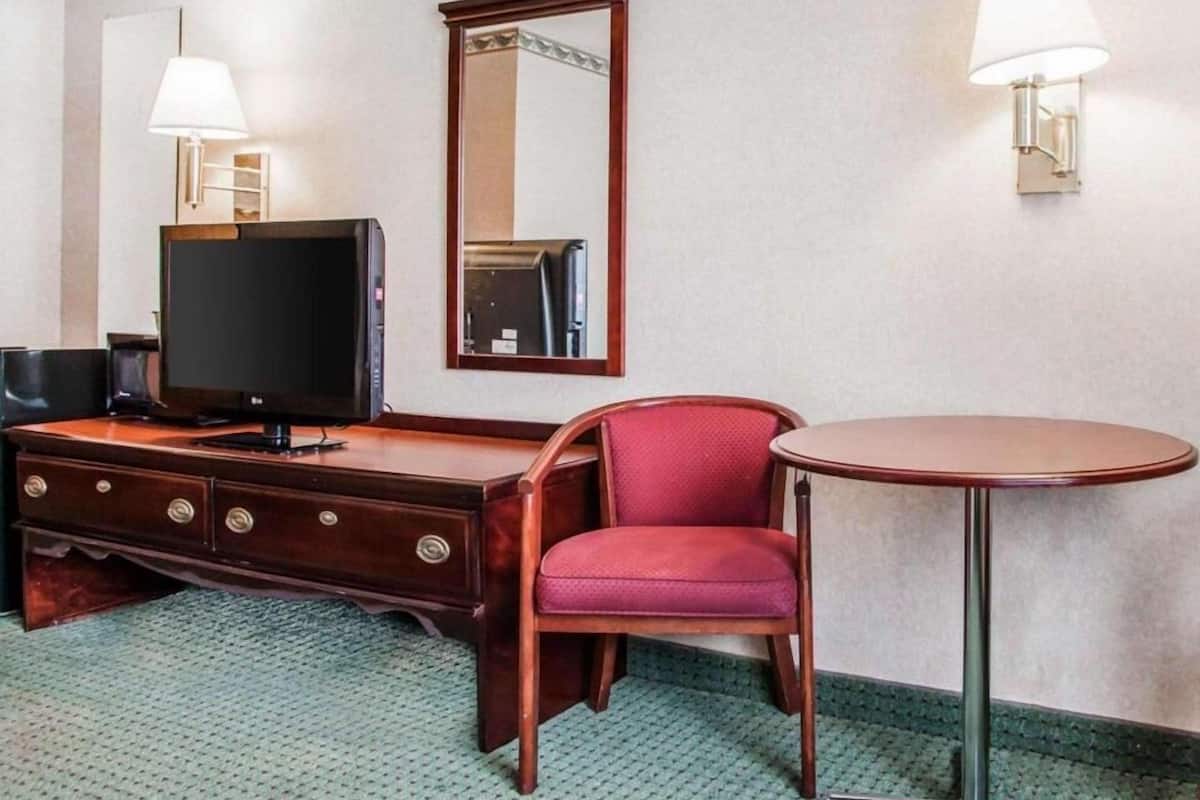 A hotel room with a wooden dresser, a television, a red chair, a round table, and two wall-mounted lamps.
