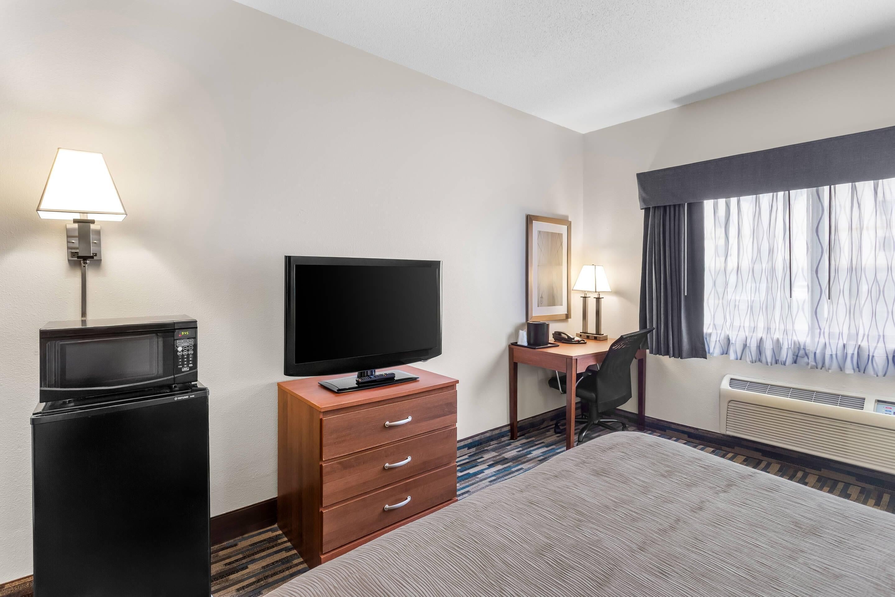 Ein Hotelzimmer mit Bett, Schreibtisch mit Stuhl, Fernseher, Mikrowelle, Kommode und einem Fenster mit Vorhängen.