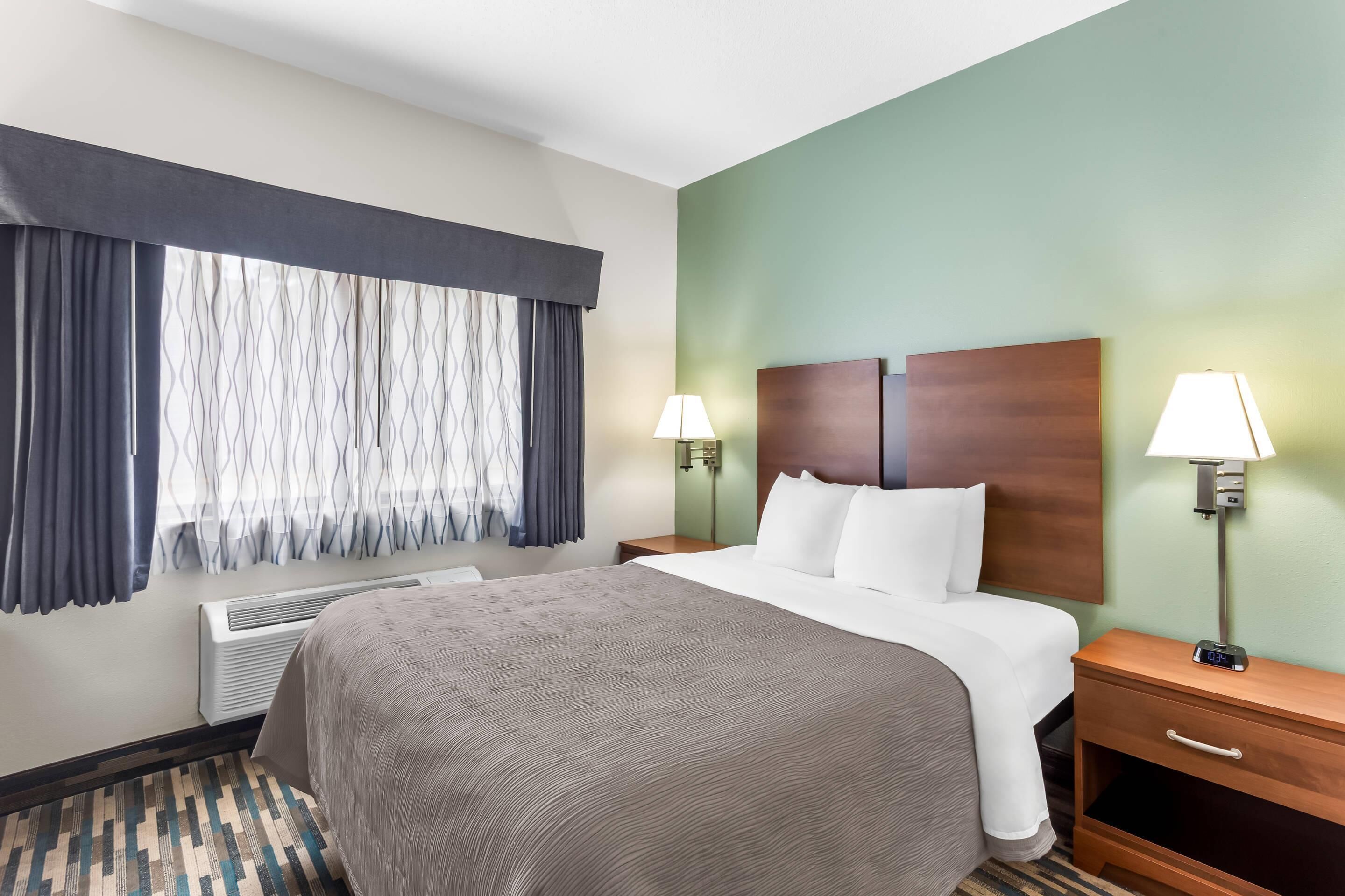 A hotel room with a large bed, two bedside tables with lamps, a heater, and a window with curtains.