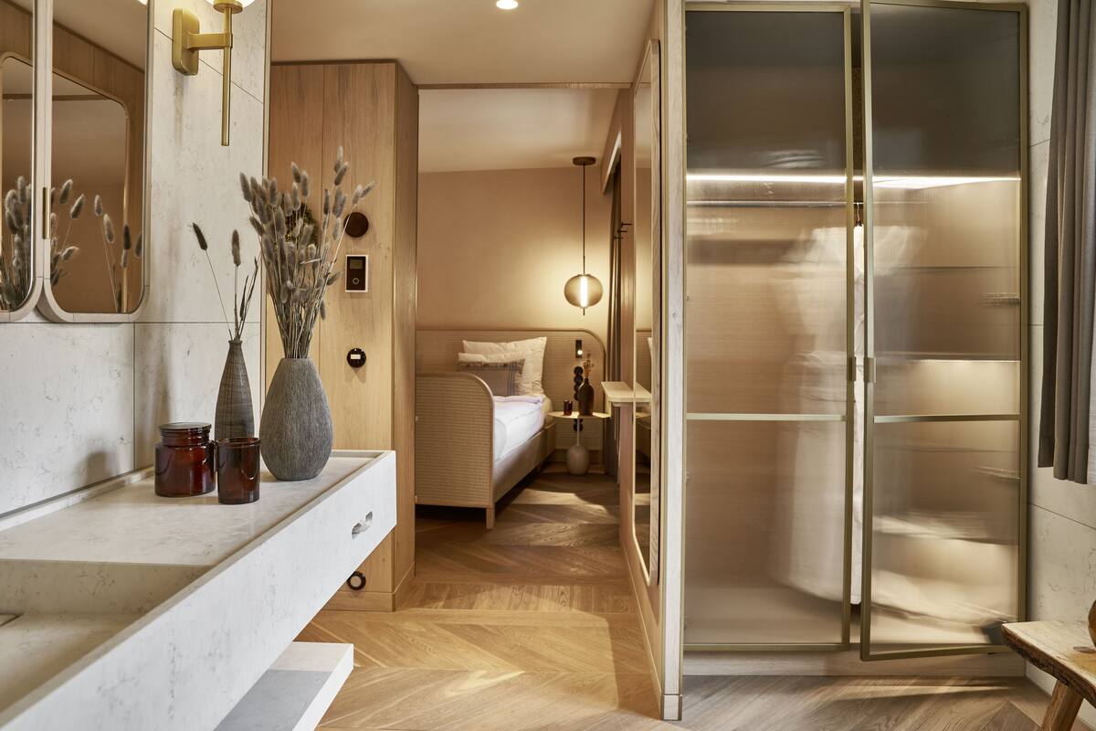 Una habitación de hotel moderna con piso de madera, un espejo grande, una mesita de noche con lámpara y un armario con puerta corredera.