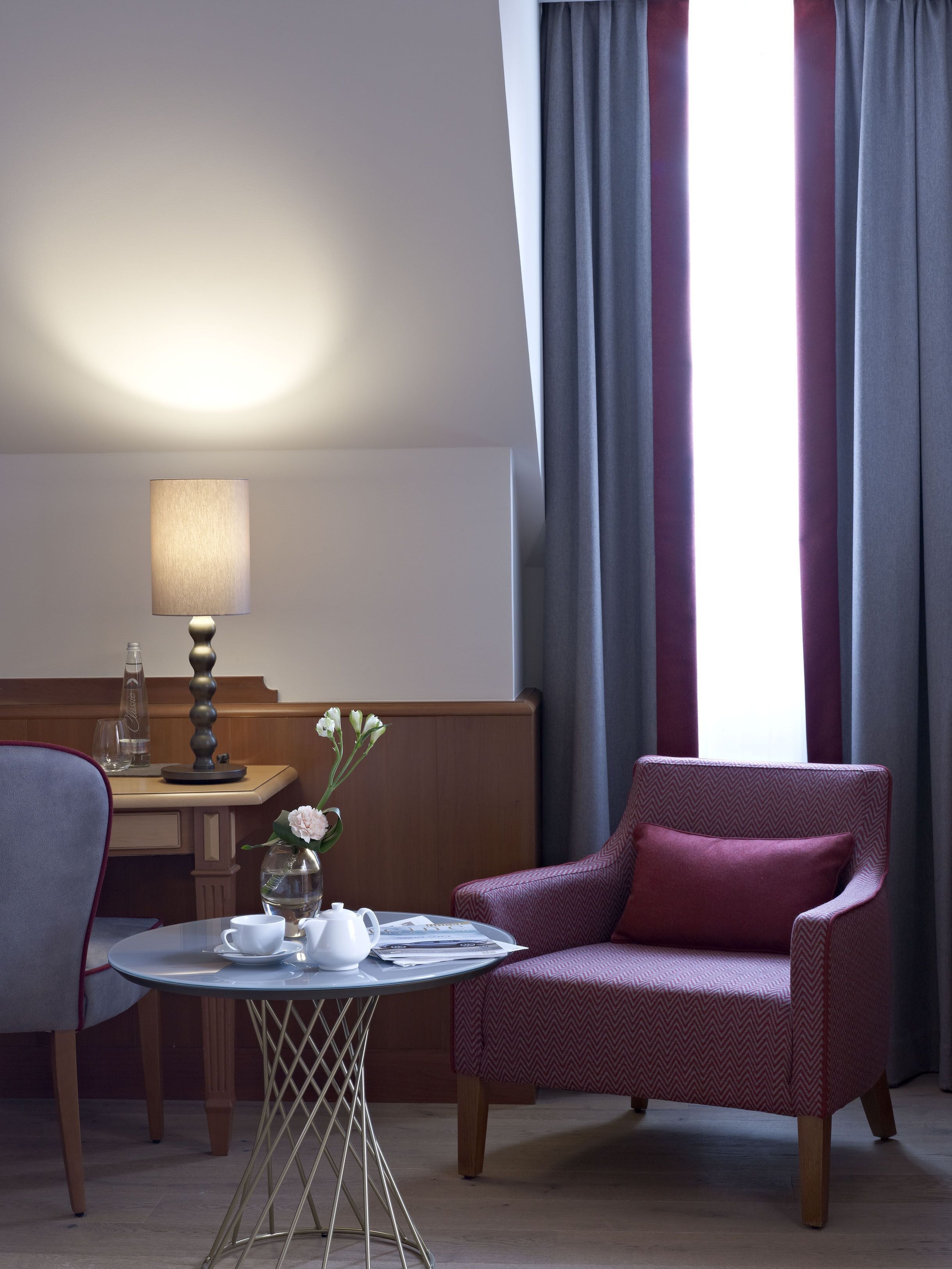 A hotel room with a round table, a chair, a lamp, and a window with curtains.