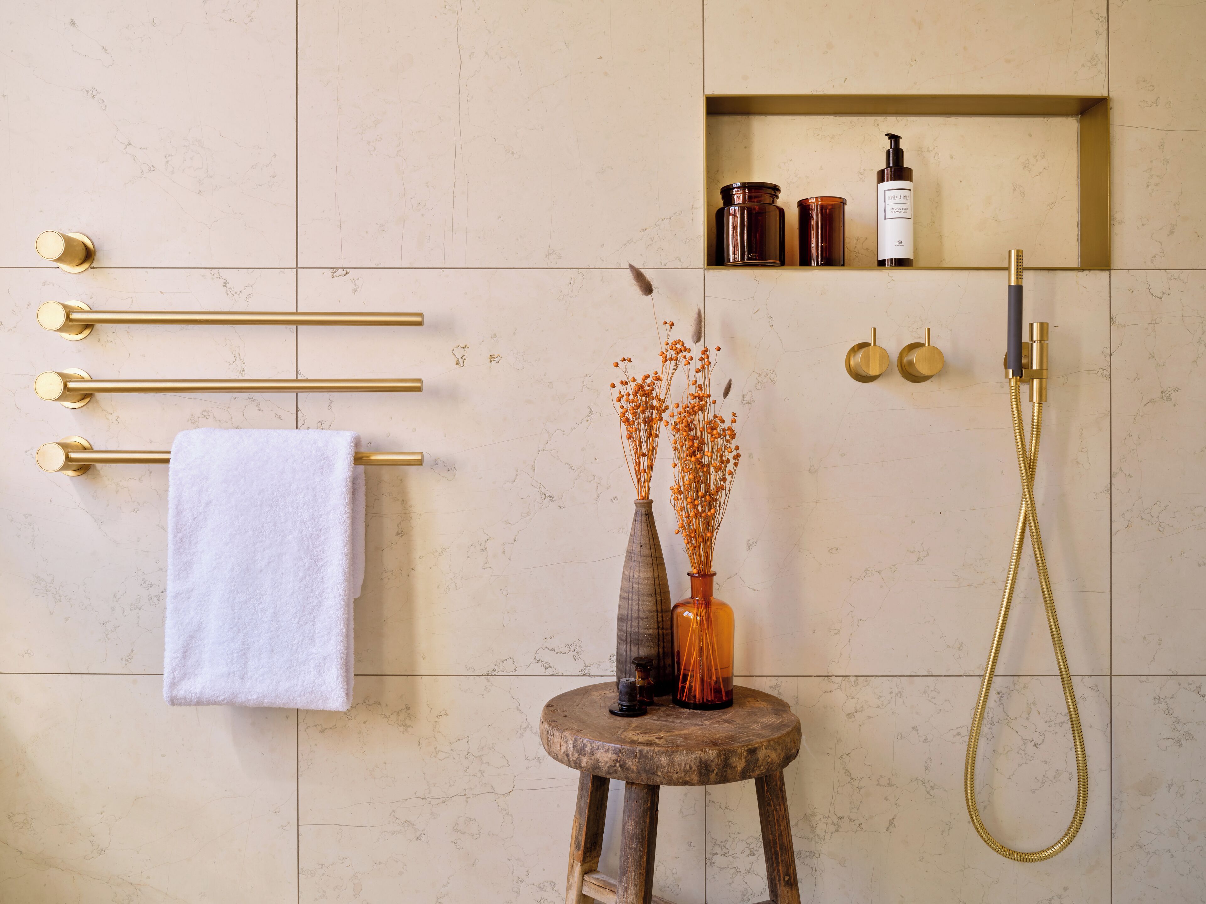 A bathroom with a marble wall, a towel rack with a white towel, a wooden stool with a vase, and a shower with a gold hose.