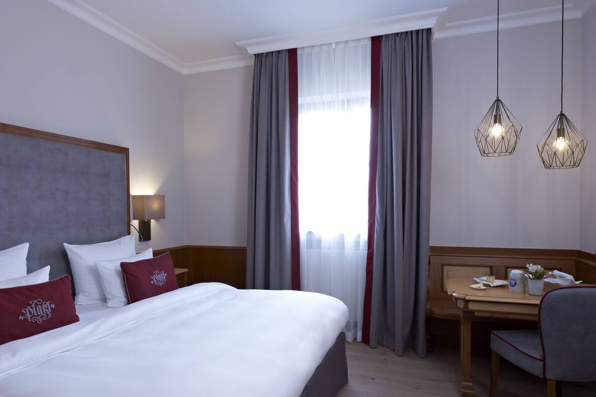 Habitación de hotel con una cama grande, un escritorio y una silla. Cuenta con una ventana con cortinas, una lámpara y dos lámparas colgantes.