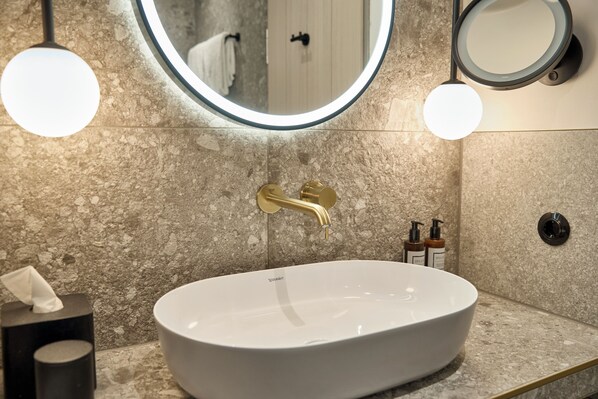 Un baño moderno con un lavamanos blanco ovalado, grifo dorado y dos espejos redondos.