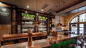A bar area with copper fixtures, a green tiled backsplash, and shelves with glassware and plants. - Platzl Hotel (Munich)