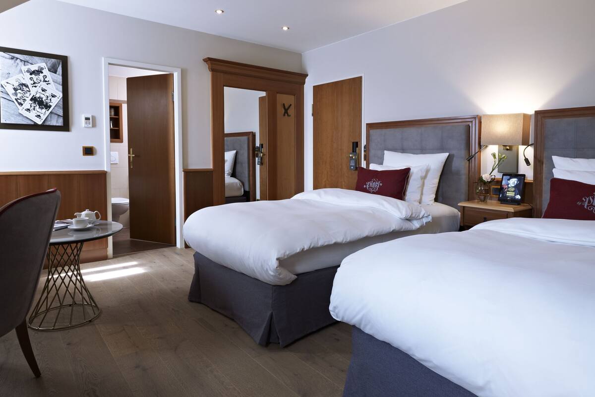 Habitación de hotel con dos camas, una mesita pequeña con una silla, una mesita de noche con una lámpara y un reloj en la pared.