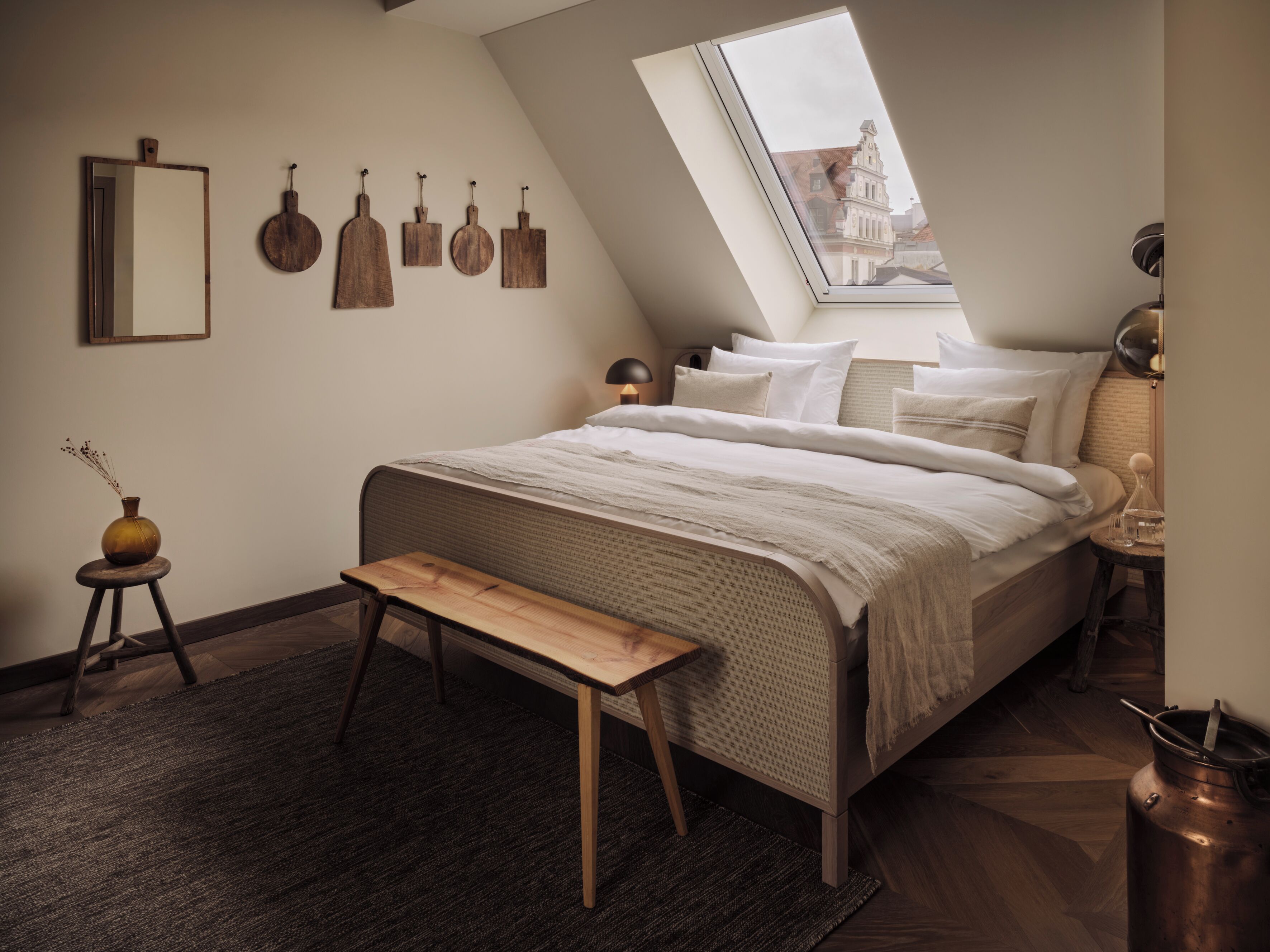Una camera da letto con soffitto spiovente, una finestra a lucernario, un letto con biancheria da letto bianca, un comodino con una lampada e una panca in legno.