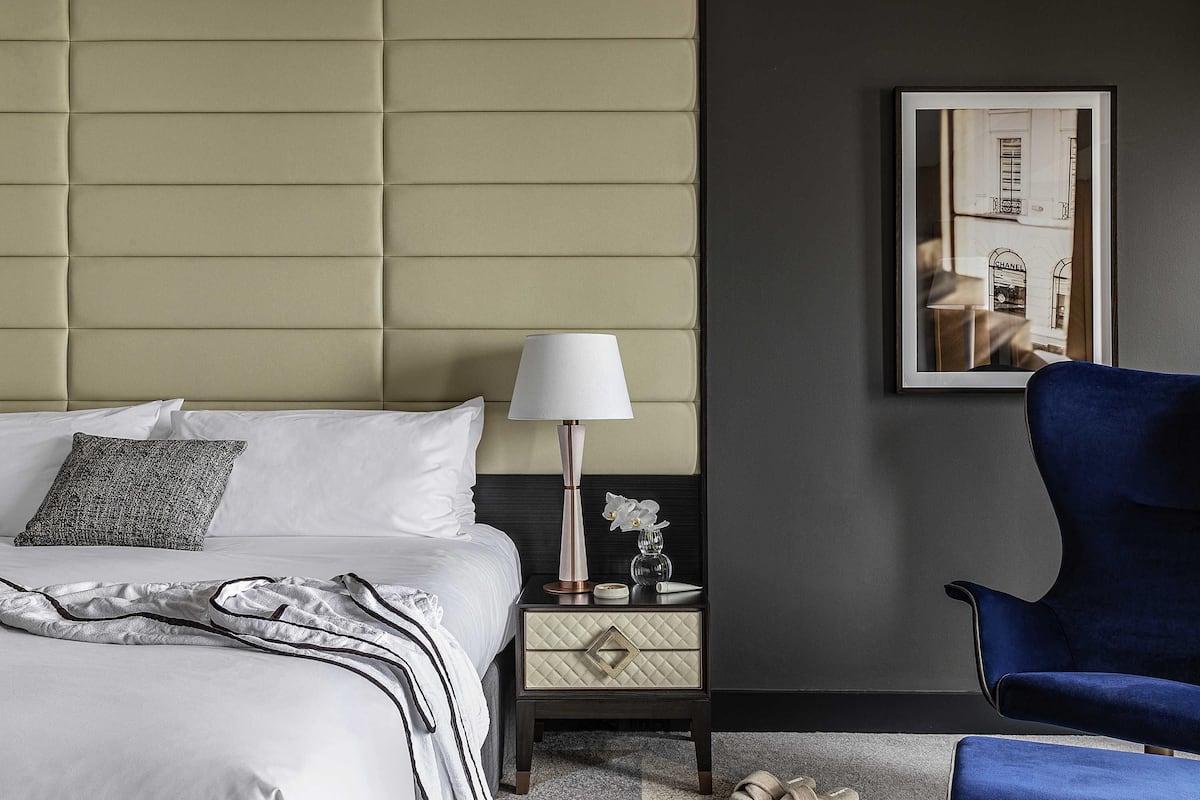 A neatly made bed with white linens and a grey pillow, a bedside table with a lamp and a small vase, a framed picture on the wall, and a blue chair.