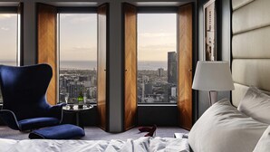 Quarto de hotel com uma cama grande, uma poltrona azul, uma mesinha com uma garrafa e um copo, e vista da cidade através de amplas janelas.