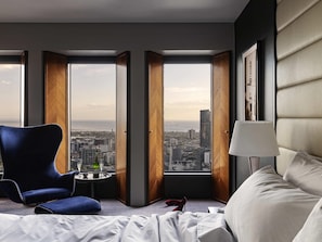 A hotel room with a large bed, a blue armchair, a small table with a bottle and glass, and a view of the city through large windows. - Sofitel Melbourne On Collins (Melbourne)