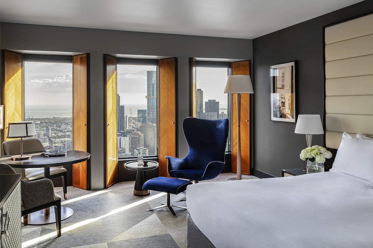 A hotel room with a large bed, a blue armchair, a desk with a lamp, and a view of the cityscape through large windows.