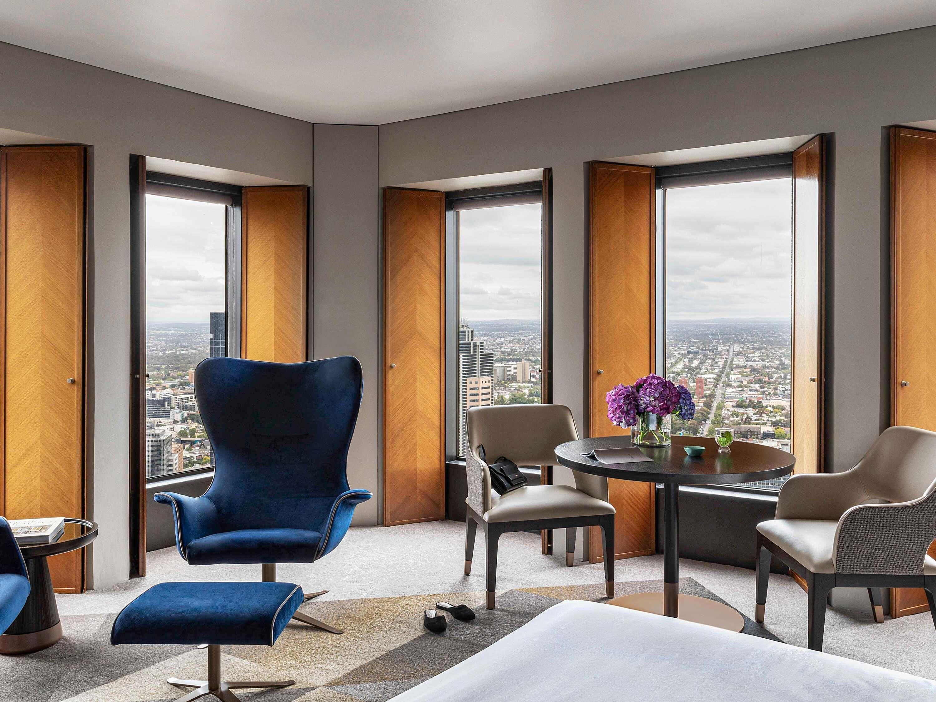 A modern hotel room with a large window, a blue armchair, a round table with a vase of flowers, and a view of the cityscape.