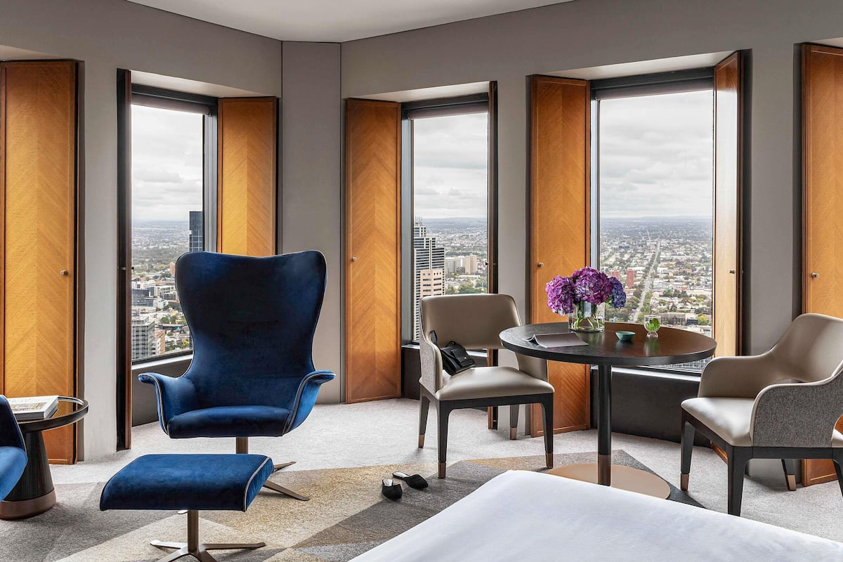 A modern hotel room with a large window, a blue armchair, a round table with a vase of flowers, and a view of the cityscape.