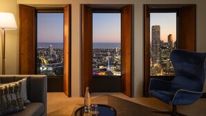Quarto de hotel com vista para a cidade, um sofá, uma cadeira e uma pequena mesa com garrafa e copo.
