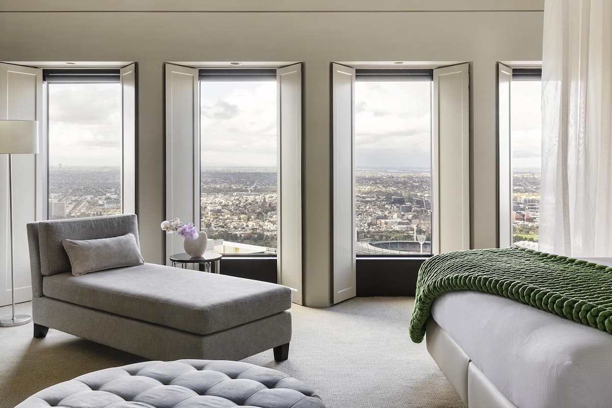 A modern hotel room with a grey sofa, a green throw, and large windows offering city views.